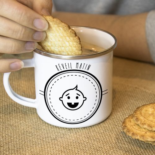 Une main tremps un biscuit dans un mug en métal émaillé avec un bébé